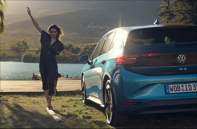 Woman dancing near a blue Volkswagen ID.3 by a lake.
