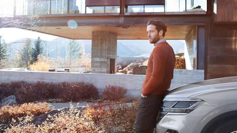 Mann lehnt an Motorhaube eines parkenden VW Touareg vor einem modernen Haus im Freien. 