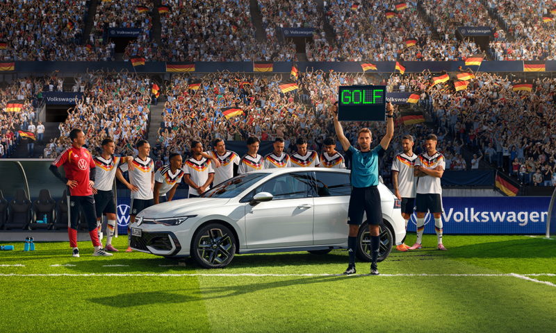 Ein weißer Golf R-Line steht mit der Herren  Fußballnationalmannschaft auf einer Rasenfläche im Stadion. Der Schiedsrichter hält die Wechselanzeige mit dem Schriftzug „Golf“ in die Höhe.​  ​