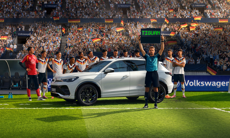 Ein weißer Tiguan R-Line steht mit der Herren  Fußballnationalmannschaft auf einer Rasenfläche im Stadion. Der Schiedsrichter hält die Wechselanzeige mit dem Schriftzug „Tiguan“ in die Höhe.​