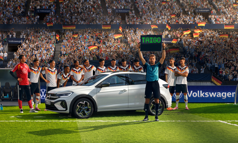 Ein weißer Taigo R-Line steht mit der Herren  Fußballnationalmannschaft auf einer Rasenfläche im Stadion. Der Schiedsrichter hält die Wechselanzeige mit dem Schriftzug „Taigo“ in die Höhe.​