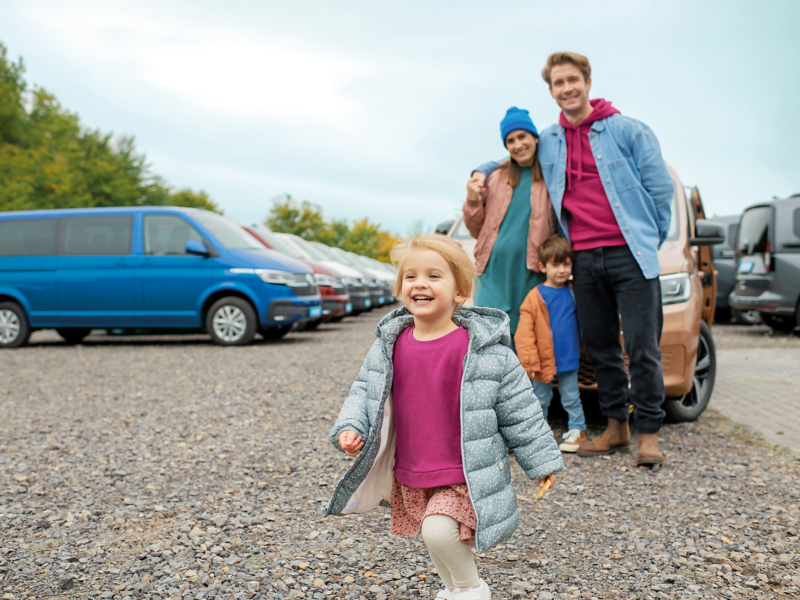 Eine Familie auf einem Autohof, im Hintergrund stehen diverse VW Fahrzeuge auf einem Schotterplatz