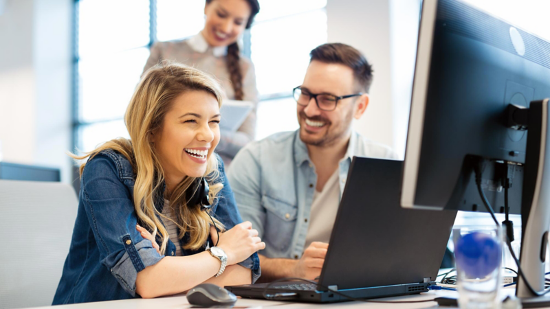 A young woman and a young man are sitting in an office, laughing in front of two computer screens