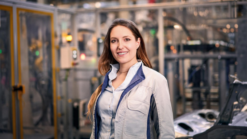A young woman wearing a Volkswagen jacket stands in a production area in front of technical equipment