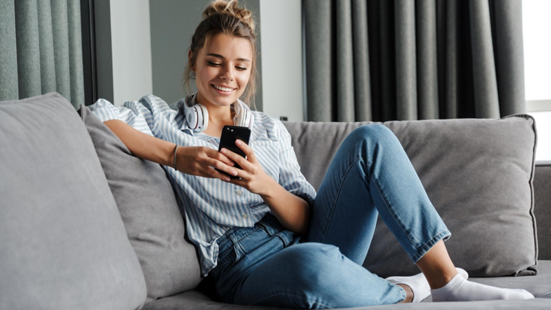A young woman is sitting on a grey sofa and looking at her phone
