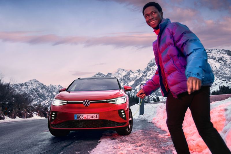 Ein junger Mann lädt seinen Volkswagen ID.4 GTX an einer Ladestation am Rand einer Straße. Die Landschaft ist winterlich.
