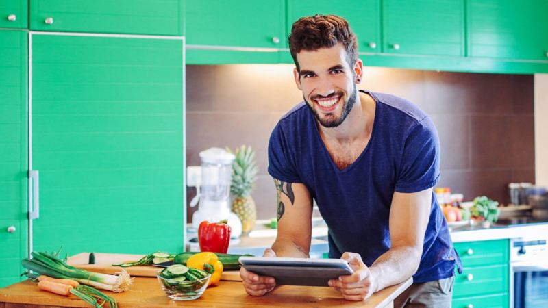 Junger Mannteht steht in einer Küche an einer Arbeitsplatte mit Gemüse und hält ein Tablet in den Händen