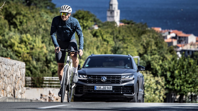 Radsportler fährt eine steile Küstenstraße hinauf, während ein Volkswagen Touareg hinter dem Fahrradfahrer folgt; im Hintergrund mediterrane Stadtlandschaft mit Meerblick und historischem Kirchturm.