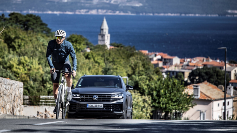 Fahrradfahrer fährt vor einem VW Touareg einen Berg hoch, im Hintergrund Stadt und Meer.