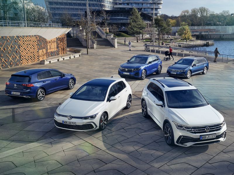 Cinque modelli Volkswagen fermi in un parcheggio. Sullo sfondo uno spazio cittadino.