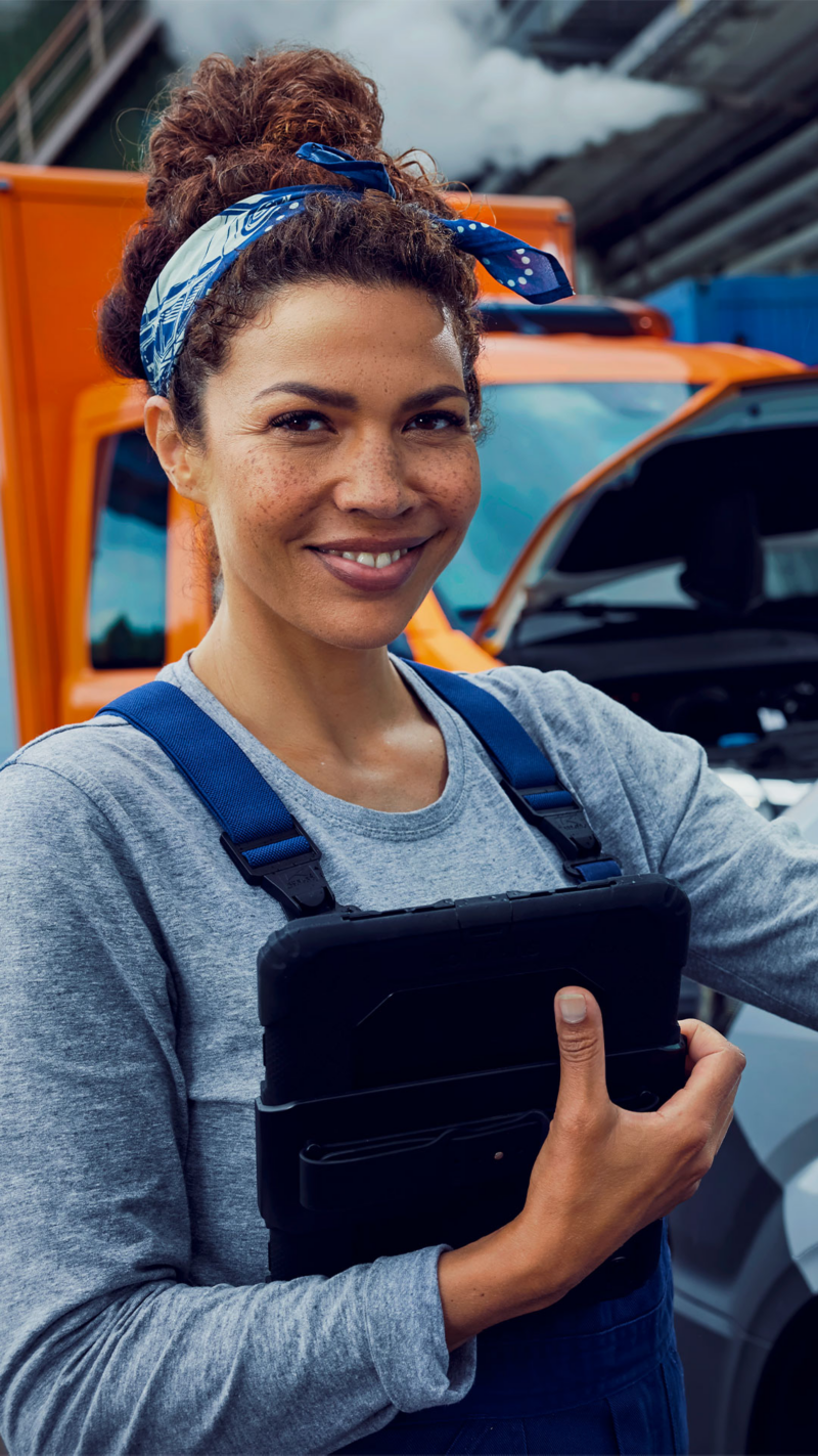 Une femme souriante en salopette bleue et bandeau, tenant une tablette, devant un véhicule orange.
