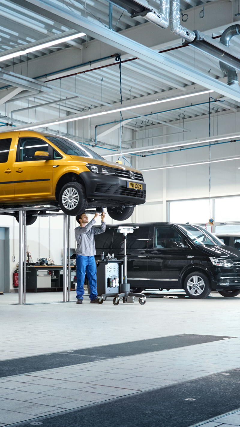  Un mécanicien travaille sous une voiture jaune soulevée dans un garage, tandis qu'une fourgonnette noire est garée à côté.