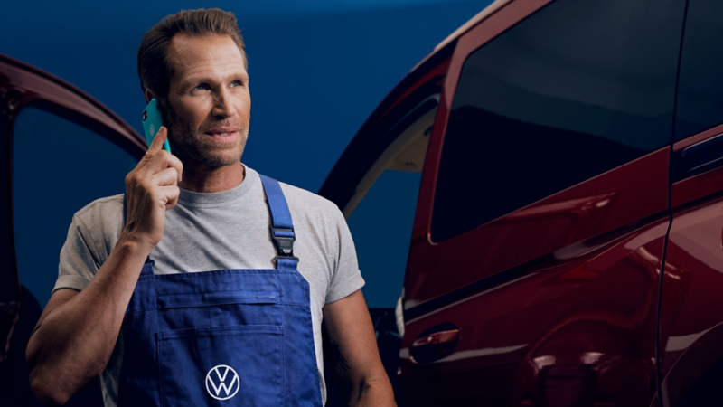  Un homme en salopette bleue avec un logo Volkswagen parle au téléphone devant un véhicule rouge.