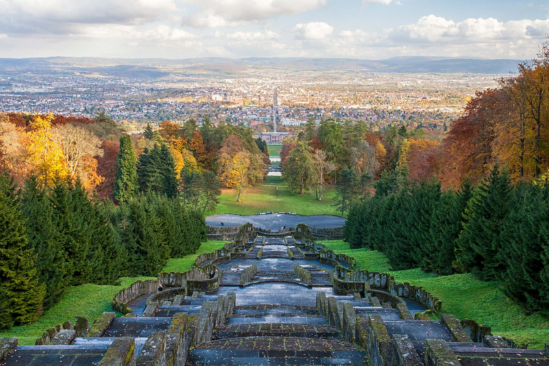 Blick vom Bergpark Wilhelmshöhe vorbei an bunten Laubbäumen auf die Stadt Kassel im Dämmerlicht