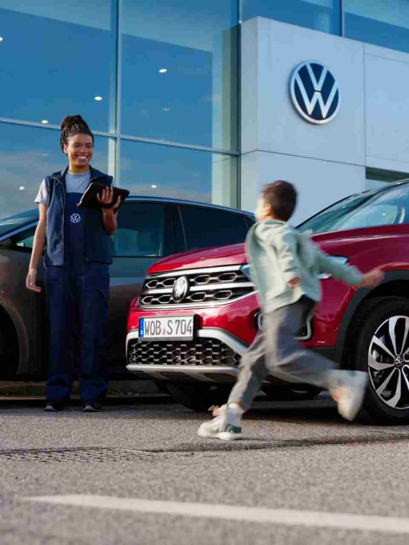 A boy runs between a bronze electric car and a red SUV at a Volkswagen dealership, while a female employee smiles with a tablet and two female customers stroll past.