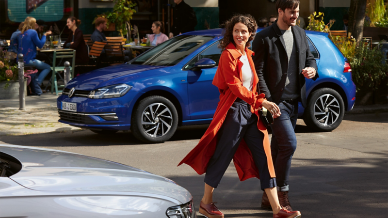 Une femme et un homme traversent une rue. En arrière-plan, une Golf VW bleue devant un café.