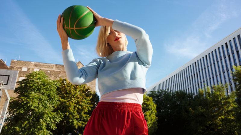 Frau springt mit Basketball in der Hand hoch. Urbanes Setting mit Stadt und Parkanlage im Hintergund. Lifestyle.