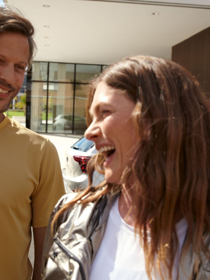 Ein lachendes Paar Mann und Frau lachen,  vor einem im Hintergrund ein parkendens MOVE Sondermodell von VW in silber