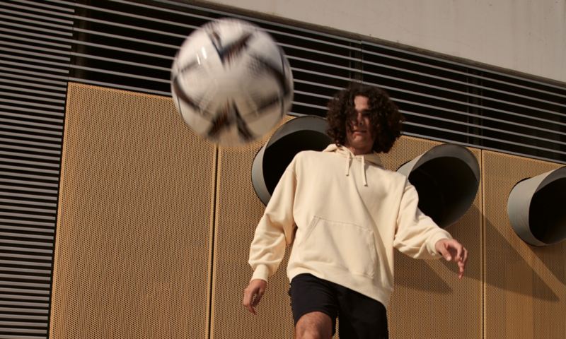 Ein Mann in kurzen Trainingshosen spielt Fußball, Ball fliegt zum Betrachtenden