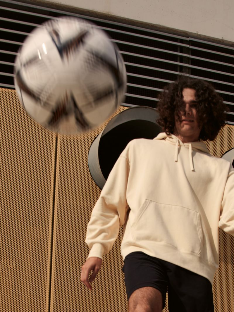 Ein Mann in kurzen Trainingshosen spielt Fußball, Ball fliegt zum Betrachtenden