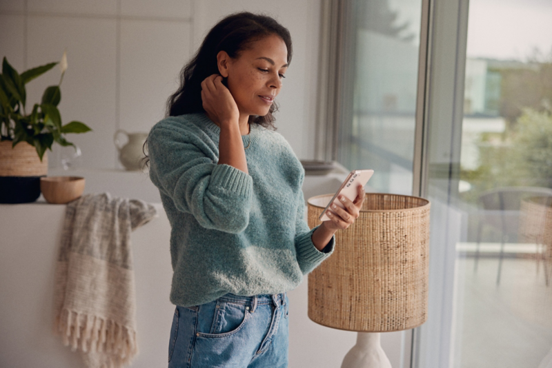 Une femme aux cheveux longs et au pull turquoise se tient à la fenêtre et reçoit des informations sur Volkswagen sur son smartphone.