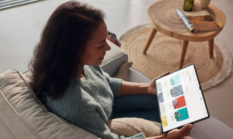 Eine Frau sitzt auf einem Sofa und schaut auf ein Tablet. Auf dem Display: die Modell-Übersicht von Volkswagen.
