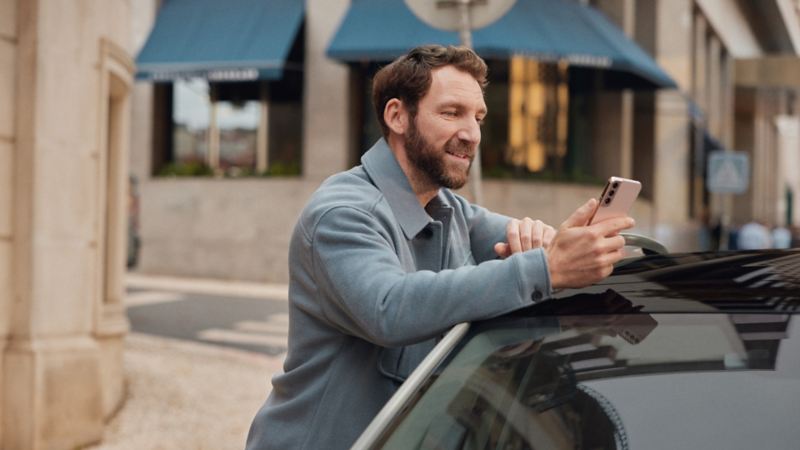 Vyras stovi atsirėmęs į „Volkswagen“ automobilį ir žiūri į savo mobilųjį telefoną