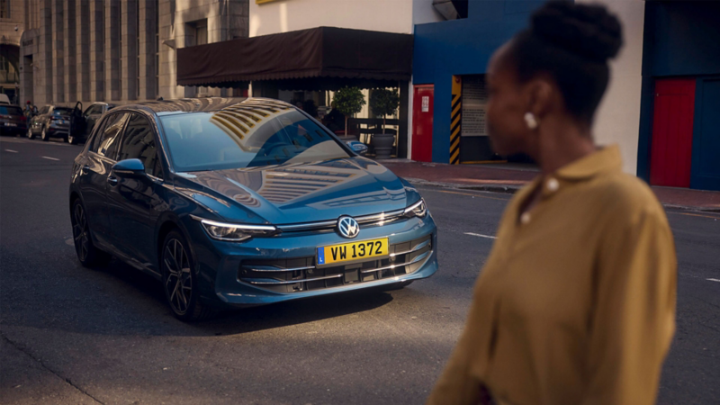 Une Golf bleue est garée dans la rue. Au premier plan se trouve une femme qui regarde la voiture en se détournant du spectateur.