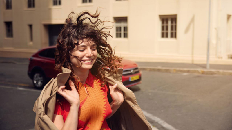 Une femme aux boucles brunes et vêtue d'un pull rouge-orange se tient devant une Volkswagen rouge.