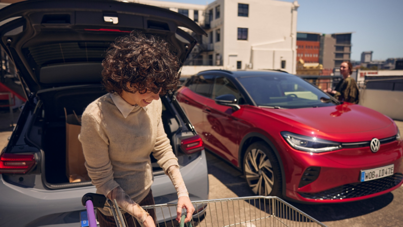 Eine Frau stellt eine Einkaufstasche in den Kofferraum eines VW ID.3 GTX, ein roter VW ID.4 GTX parkt daneben.