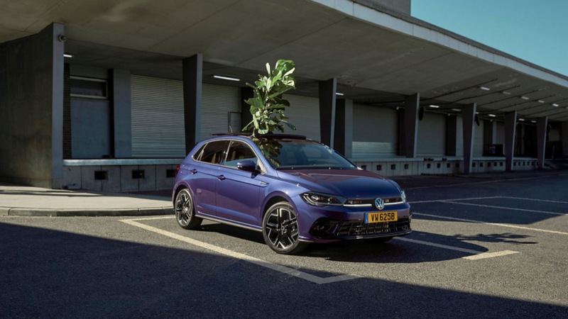 Une Polo bleue est garée sur un parking devant une zone de chargement d’un entrepôt et a chargé une plante verte à travers le toit ouvrant ouvert.