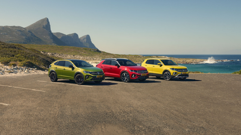 Une Taigo verte, une T-Cross rouge et une T-Roc jaune sont garées sur un parking en bord de mer, devant un panorama rocheux.