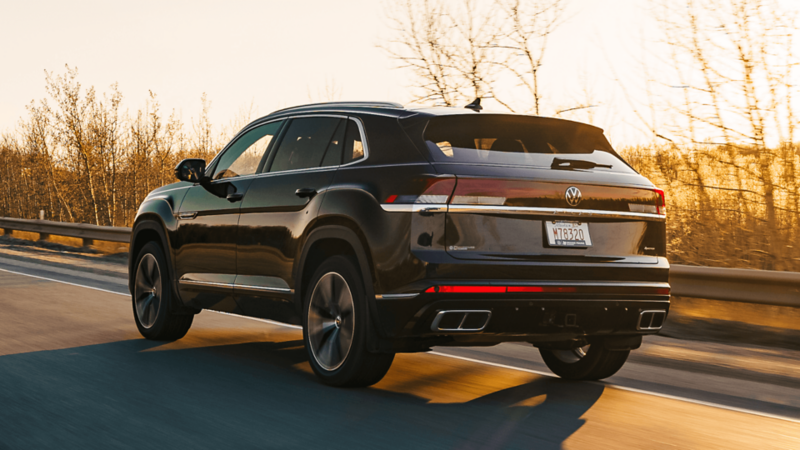 Rear-side view of a black Volkswagen Atlas Cross Sport 4MOTION driving on a highway at sunset, with golden light reflecting off the vehicle.