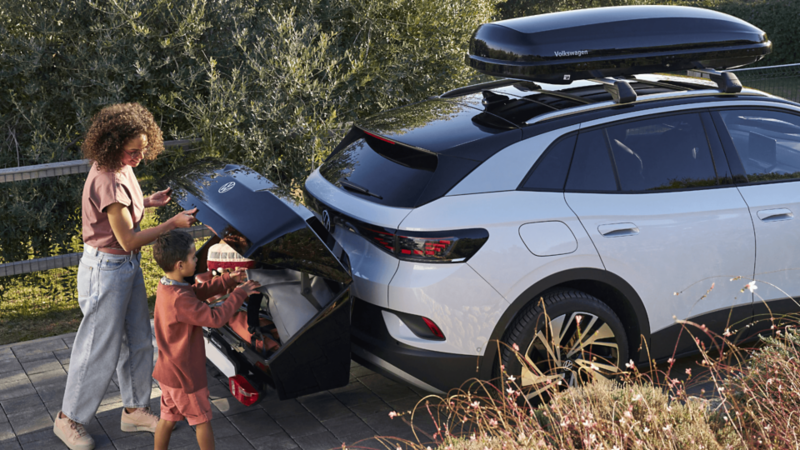 Mother and child loading luggage into the rear of a Volkswagen ID.4 SUV, equipped with a roof box, parked outdoors. 