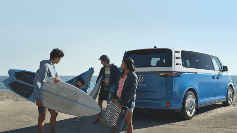 Rear view of a blue Volkswagen ID. Buzz at the beach with people and surfboards.