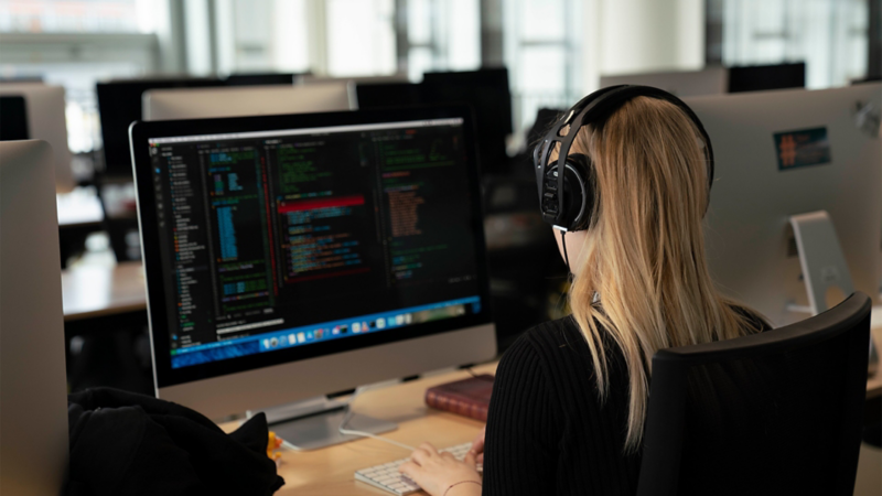 Junge Studentin bei 42 Wolfsburg sitzt mit Kopfhörern vor einem großen Computer-Bildschirm 