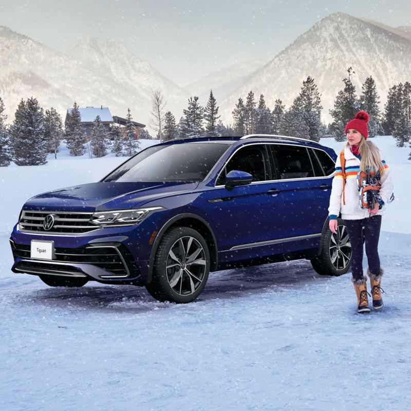 We see a woman wearing winter clothes standing by a dark blue Tiguan in a winter landscape.