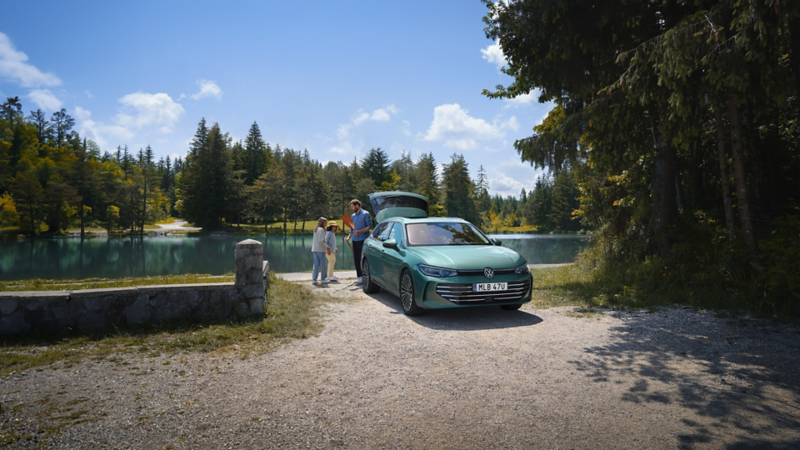 En VW Passat står parkerad vid vattnet, en man såt vid bilens bagagelucka och ger en paddel till två barn