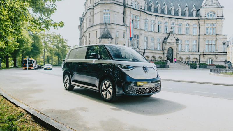 Un van électrique Volkswagen ID. Buzz bicolore (noir et blanc) est garé sur le côté d'une rue pavée, avec un bâtiment ancien et d'autres véhicules en arrière-plan.