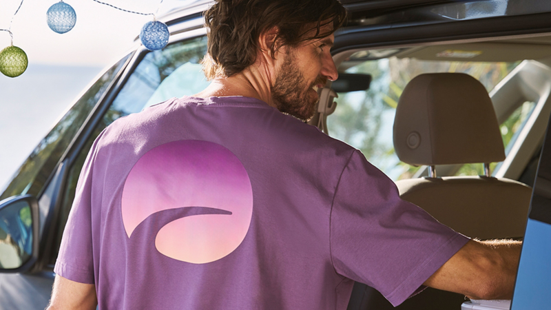 Ein lächelnder Mann mit dunklem Haar, der ein violettes T-Shirt mit einem großen, farbverlaufenden Aufdruck auf dem Rücken trägt, steigt in einen Campervan ein.