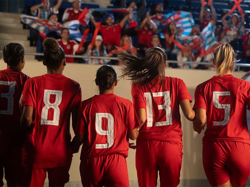 Une photographie prise de dos, capturant l'équipe de football féminine luxembourgeoise dans leurs kits rouge et blanc. Les numéros au dos de leurs maillots sont clairement visibles.