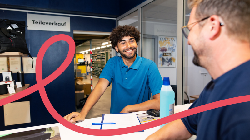 Junger Auszubildender im blauen Polohemd berät Kunden am Tresen des Teileverkaufs.