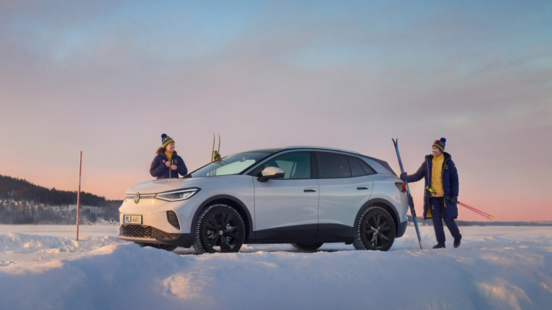 En vit ID.4 står parkerad i ett vinterlandskap. Två skidåkare med OS-kläder och skidutrustning står vid bilen