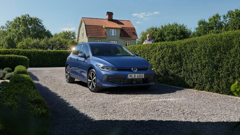 En blå VW Golf står parkerad intill en häck, en granne sneglar över häcken på bilen