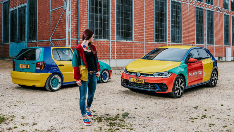 Une femme, vêtue d'une veste multicolore, se tient entre deux Volkswagen Polo aux couleurs vives et audacieuses. La Polo à l'arrière-plan est un ancien modèle tandis que la nouvelle Polo, au premier plan, est garée sur la droite. Leurs carrosseries présentent un motif de couleurs jaune, rouge et bleu.