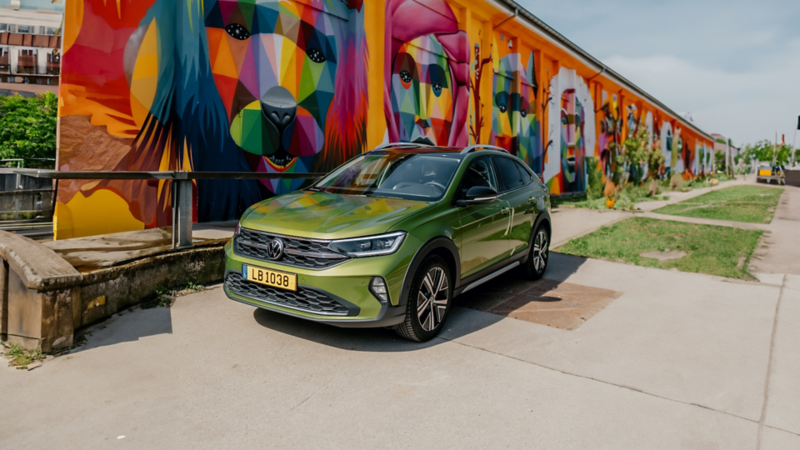 Une Volkswagen Taigo vert pomme est garée devant un grand mur de briques recouvert d'une fresque colorée et abstraite. La voiture est orientée vers l'avant, avec le soleil qui se reflète sur sa peinture.