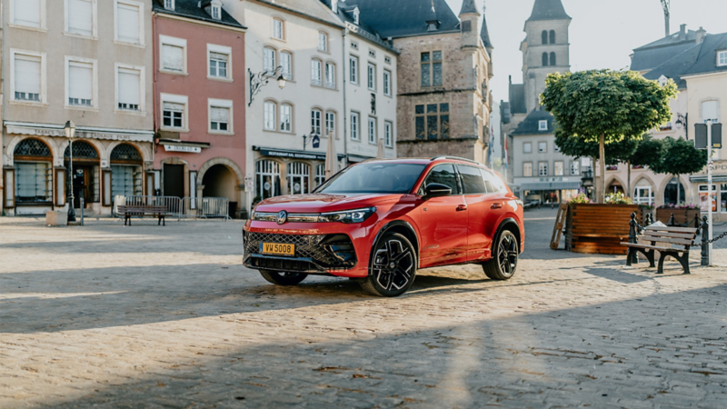  Un SUV Volkswagen Tiguan rouge est garé sur une place pavée, au milieu d'une ville historique. La voiture, garée en diagonale, est mise en évidence, avec les bâtiments et le mobilier urbain flous en arrière-plan.