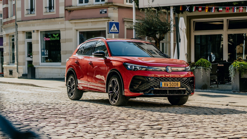 Une VW Tiguan rouge est garée au bord de la route. Des magasins sont visibles à l'arrière-plan.