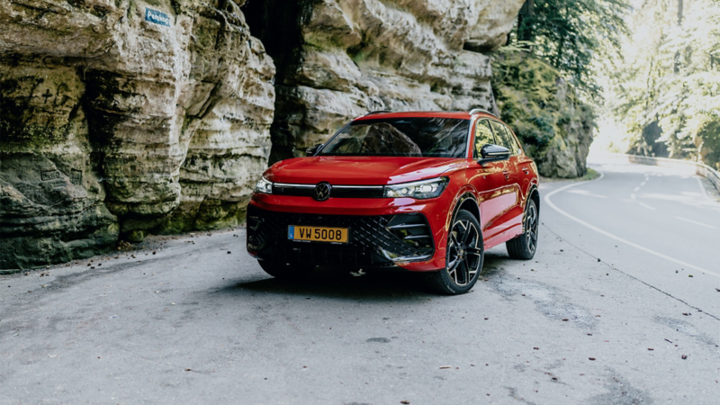  Un SUV Volkswagen Tiguan rouge est garé sur le côté d'une route sinueuse, avec des falaises rocheuses et de la végétation en arrière-plan.