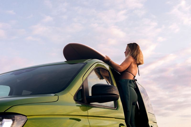 VW California de coté avec une femme qui charge une planche de surf sur le toit